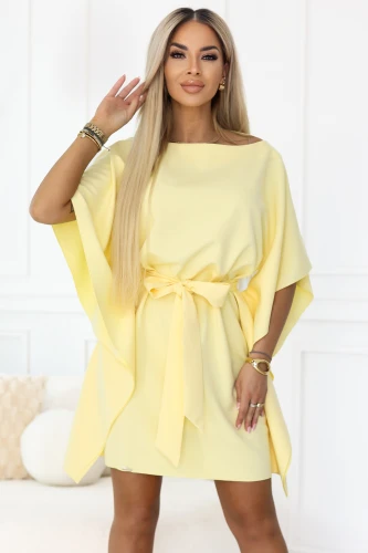 Modelka prezentuje pastelową cytrynową sukienkę mini Numoco z luźnymi rękawami typu kimono i wiązaniem w pasie, które podkreśla talię. Elegancka, lekka kreacja idealna na lato, przyjęcia i stylowe wyjścia.}