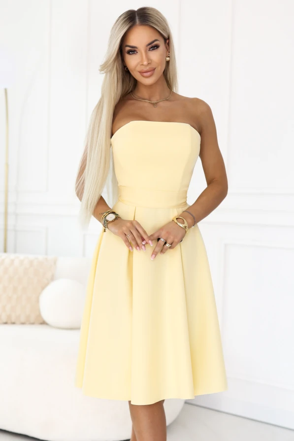 Modelka prezentuje **żółtą sukienkę midi Numoco** w kolorze **pastelowy cytrynowy**, bez ramiączek, z dopasowaną górą i rozkloszowaną spódnicą. To idealna **sukienka na wesele**, która pięknie układa się w ruchu i podkreśla talię.
