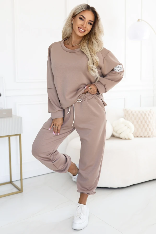 Modelka w ciemnobeżowym komplecie dresowym Numoco Basic: luźna bluza z okrągłym dekoltem i naszywką na rękawie oraz spodnie z bocznymi kieszeniami i wiązaniem w pasie.