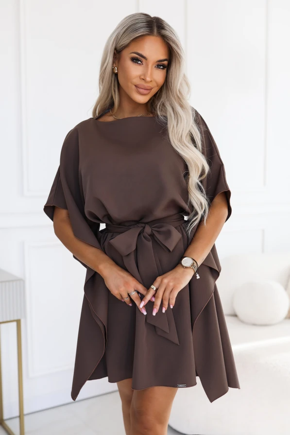 Modelka w sukience SOFIA marki Numoco w odcieniu brązu mokka. Sukienka sięga przed kolana, ma dekolt w łódkę, szerokie motylkowe rękawy i pasek zawiązany w talii na kokardę.