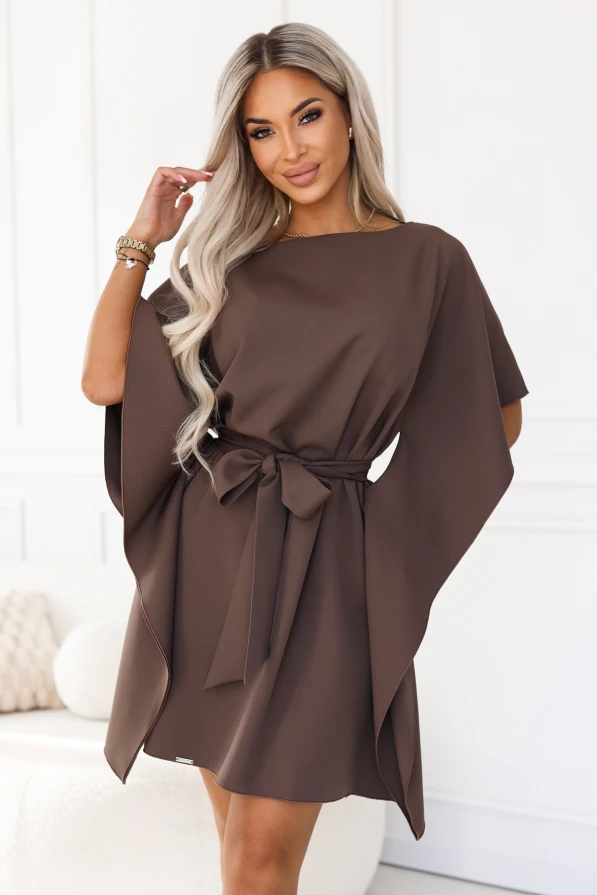 Modelka w sukience SOFIA marki Numoco w odcieniu brązu mokka. Sukienka sięga przed kolana, ma dekolt w łódkę, szerokie motylkowe rękawy i pasek zawiązany w talii na kokardę.