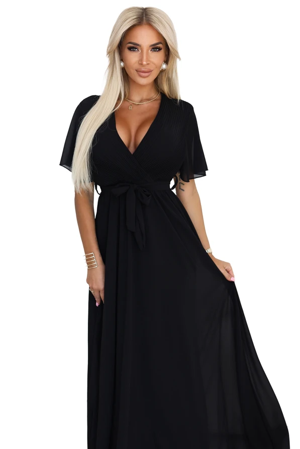 Modelka prezentuje elegancką czarną sukienkę maxi z szyfonu, z plisowanym dekoltem w kształcie litery V, krótkimi rękawkami i wiązaniem w talii; lekka i kobieca stylizacja marki Numoco Basic, idealna na wyjątkowe okazje.